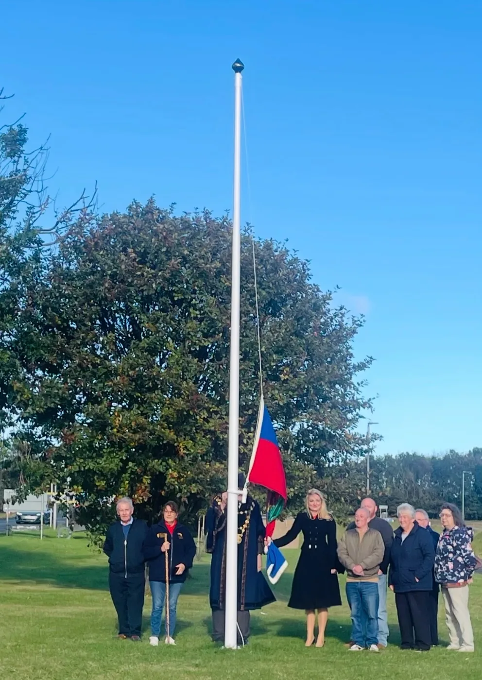The Berwick flag being raised up a white flagpole at Newfields, with a group of people standing on the grass beside it