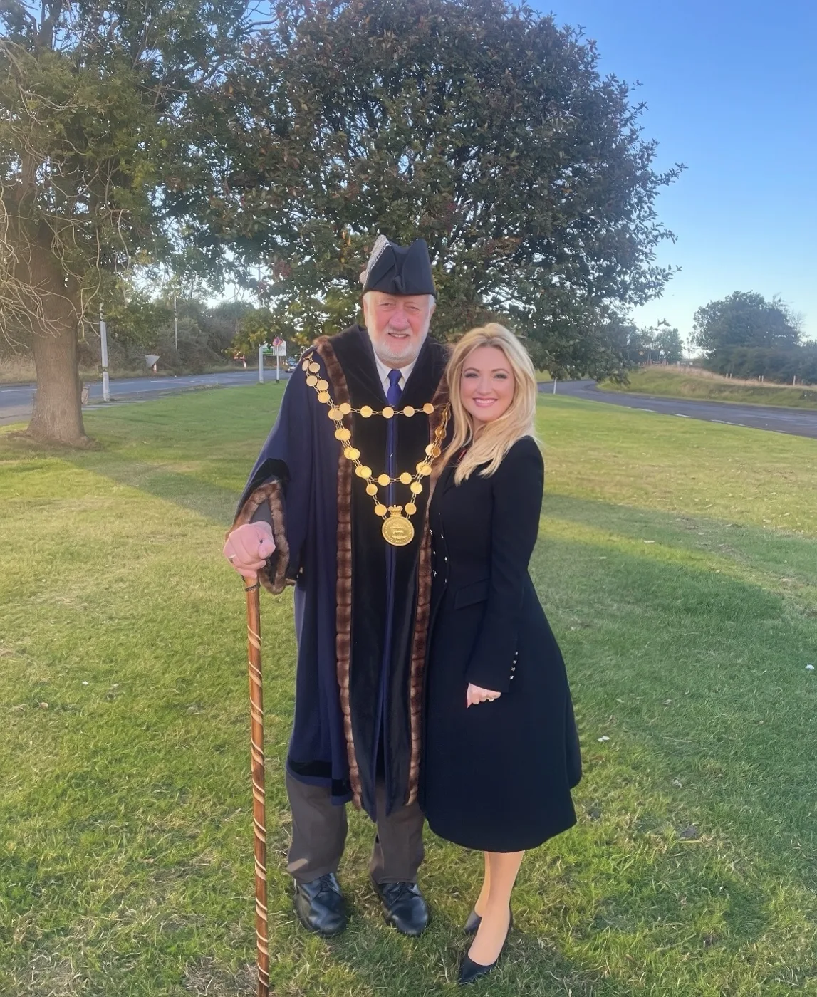 Berwick's Sheriff in a dark robe trimmed with fur, a gold chain of office and a feathered hat, holding a carved staff, standing on the grass at Newfields