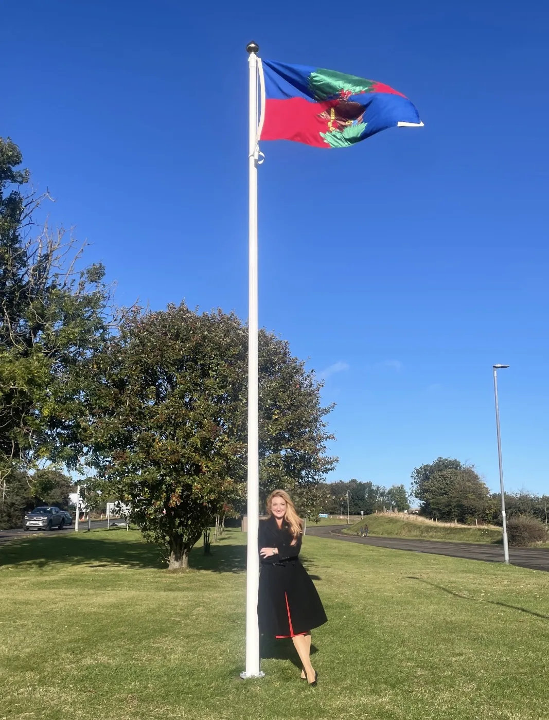 The Berwick flag flying from a white pole above a grassy verge at Newfields, with a woman in a dark coat standing beside the pole