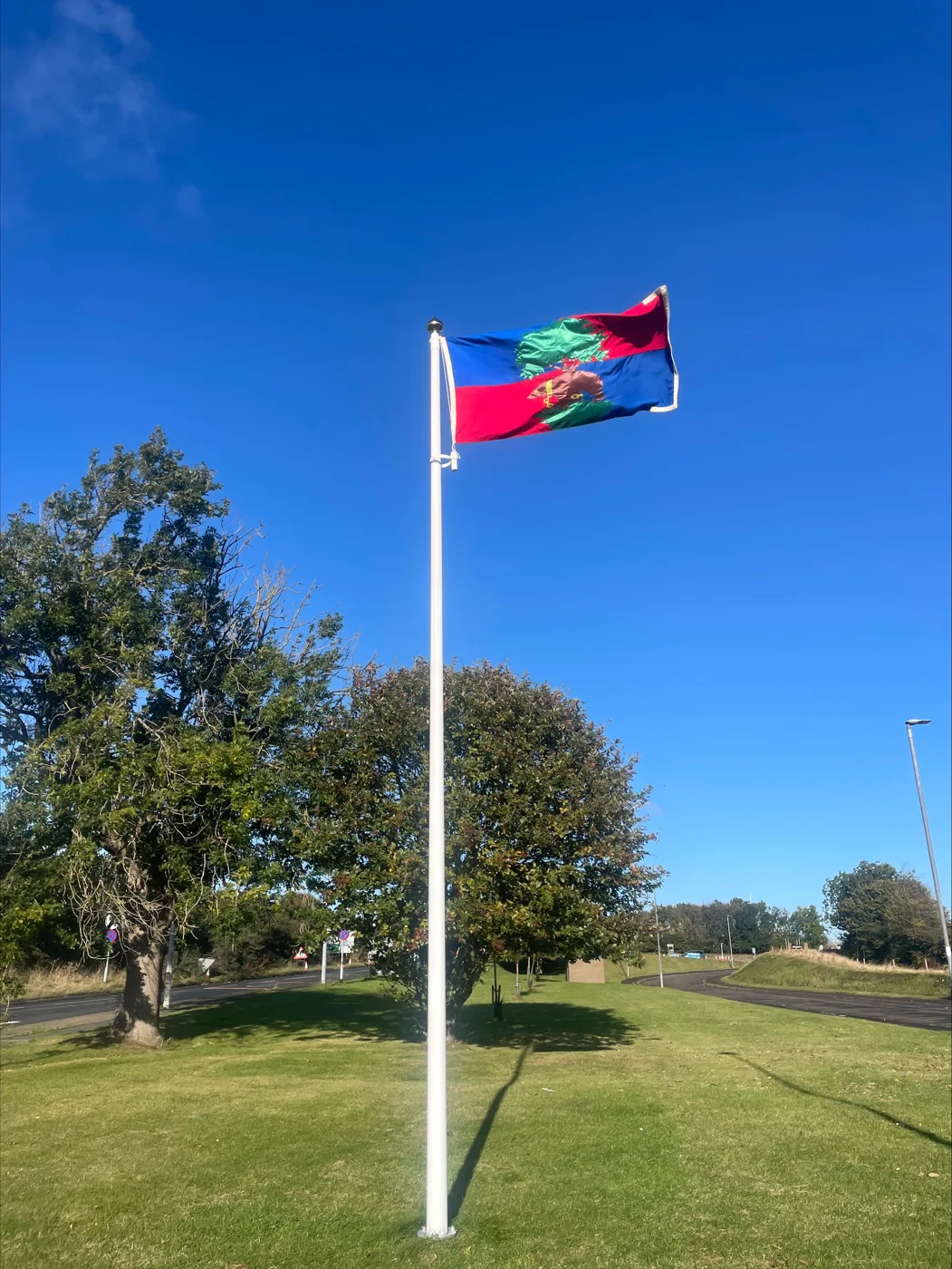 The blue and red Berwick flag with its green tree and bear emblem flying from a white pole at Newfields under a clear sky