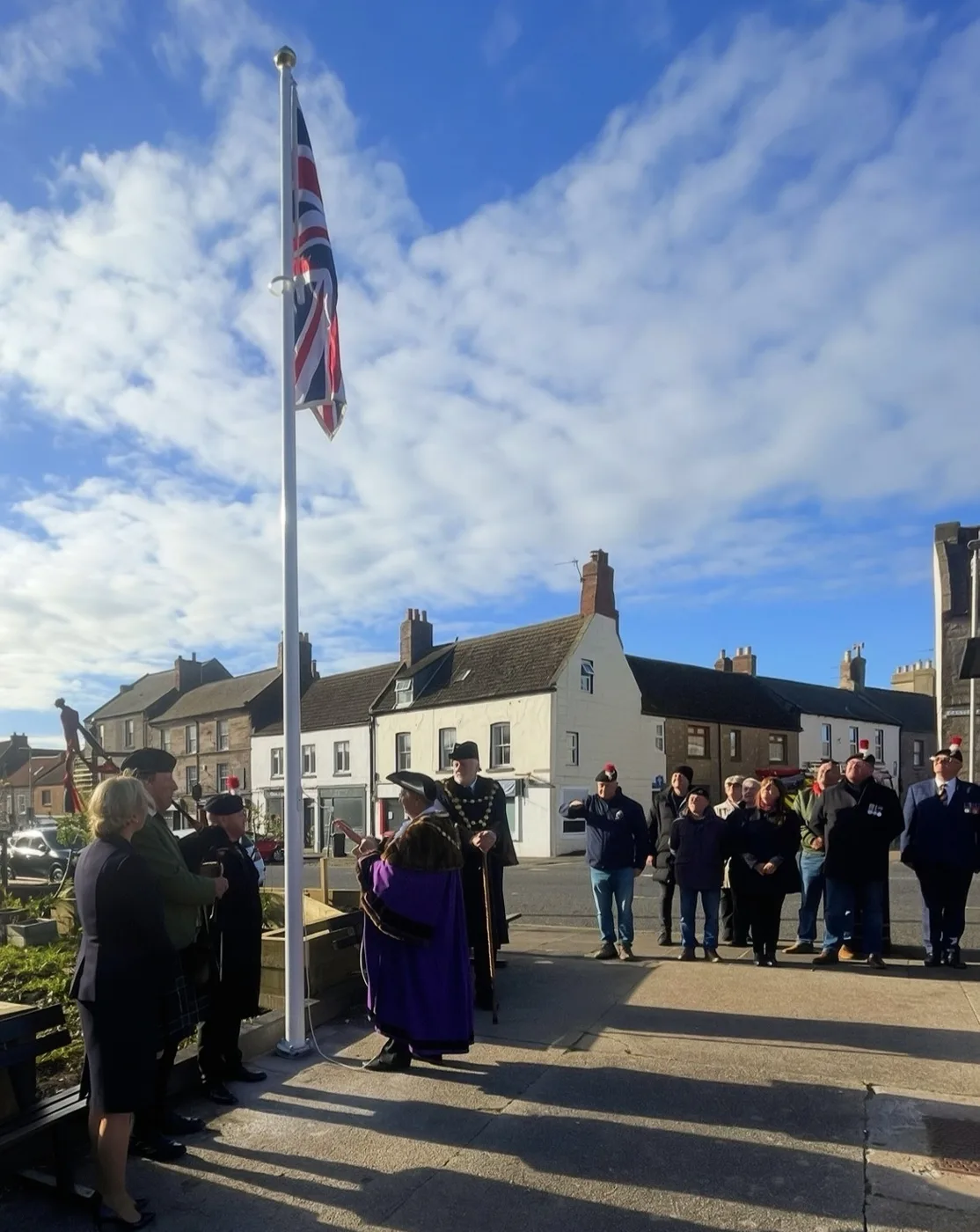 The Union Flag at the top of a tall white flagpole beside Berwick War Memorial, with a figure in purple civic robes at the foot of the pole and a line of people watching from the pavement