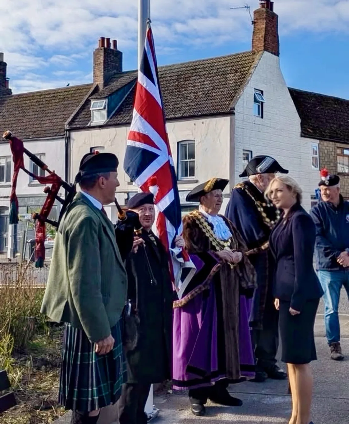 A piper in a green tweed jacket and kilt beside the Union Flag, with civic officers in robes and chains of office, veterans in berets and a woman in a dark suit