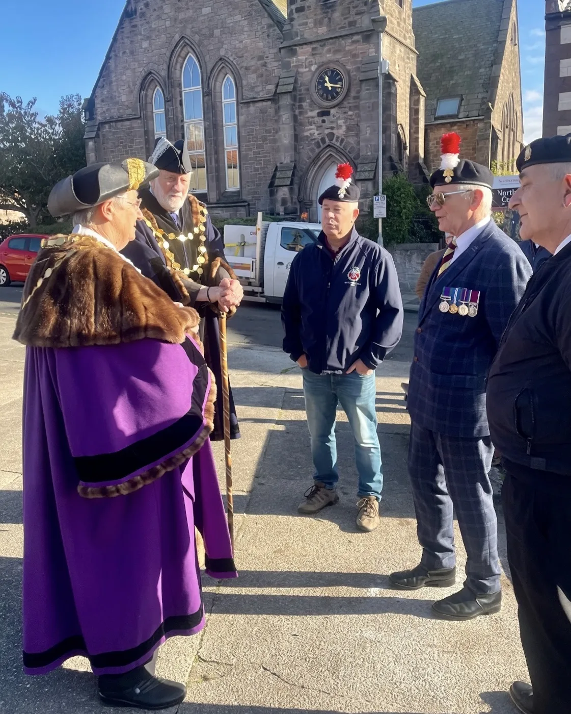Two people in civic robes, one purple and fur-trimmed, talking with men in berets and regimental hackles, one wearing medals, outside a stone church