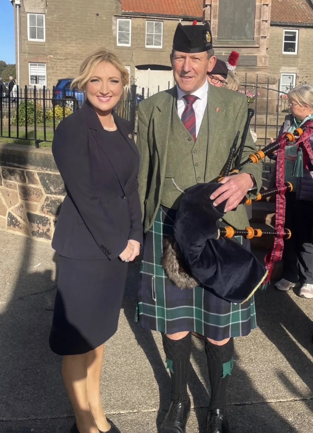 A piper in a kilt holding bagpipes standing beside a woman in a dark suit near the railings of Berwick War Memorial