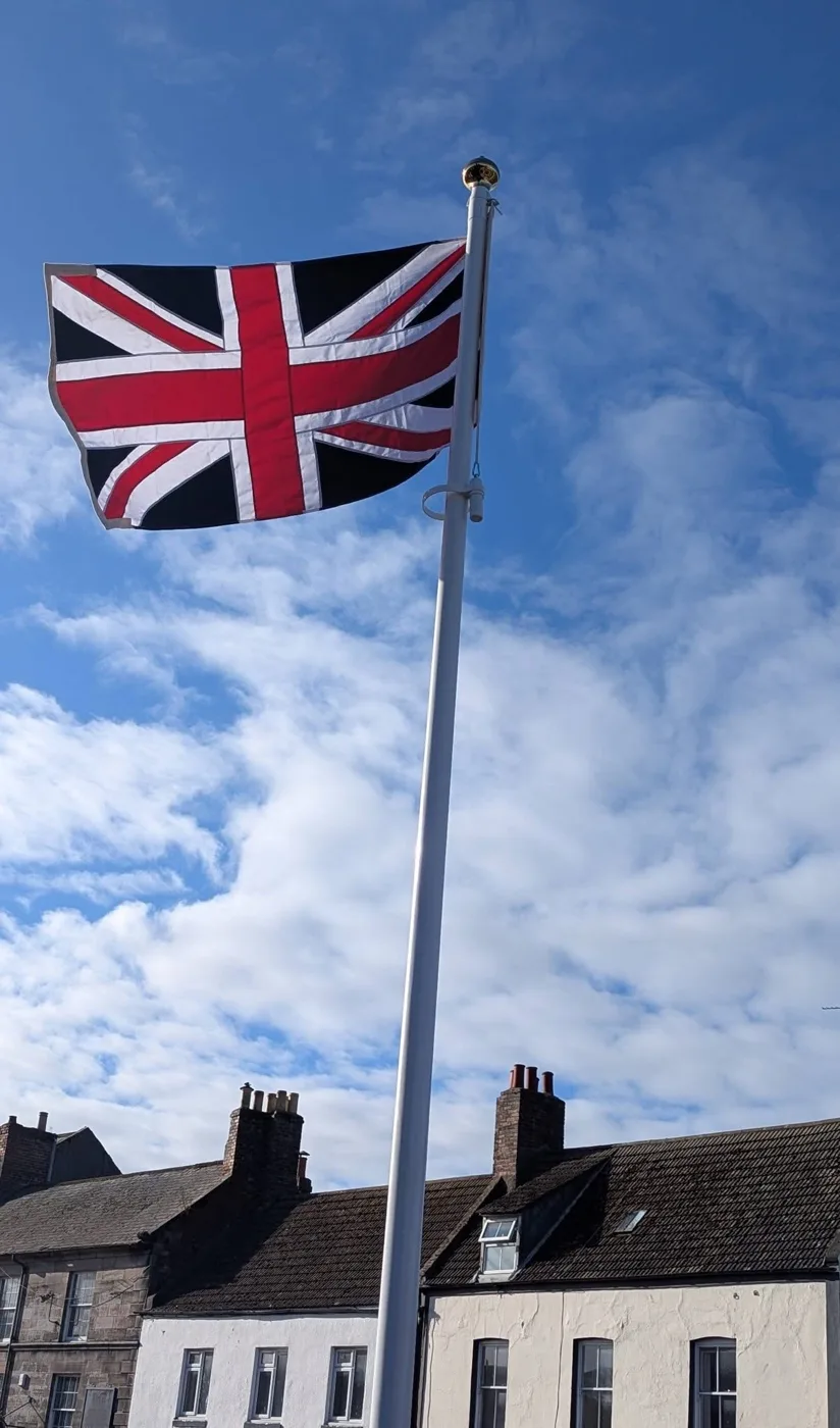 The Union Flag flying from a white flagpole with a gold finial against a blue sky and white cloud, above a row of rooftops